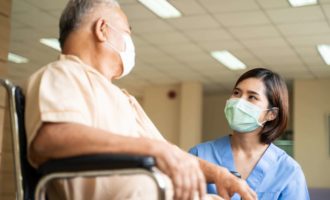 Asian nurse taking care of mature male patient sitting on wheelchair in hospital. Young woman and old man wearing surgical face mask for protection of The COVID pandemic. Girl talking to elderly man.