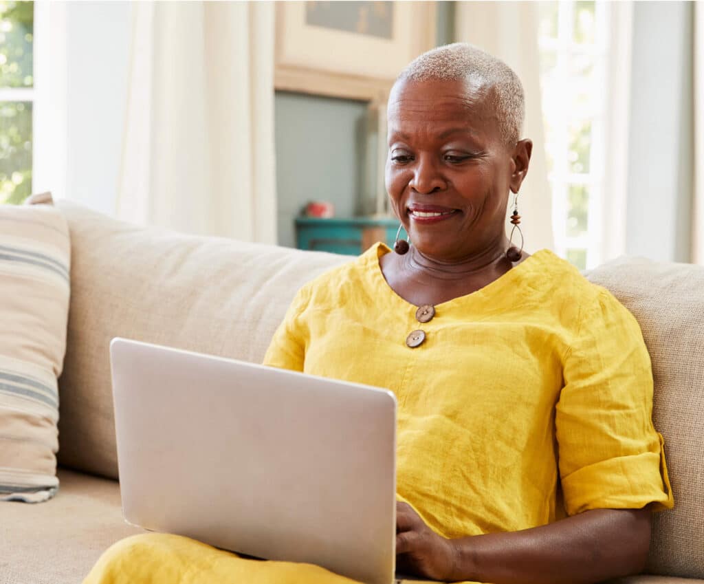 A senior woman is sitting on a couch in her living room, smiling at a tablet screen whilst participating in neurocognitive assessment, highlighting the convenience of digital platforms for cognitive health